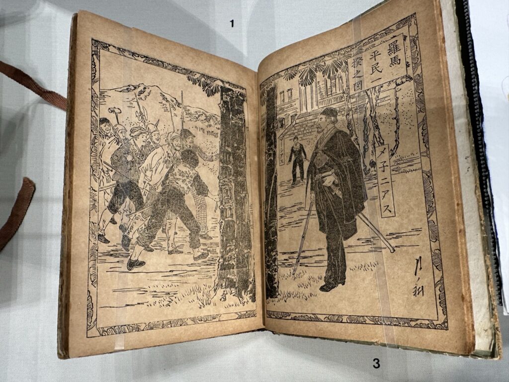 一本打开日本木刻《科利奥兰纳斯》场景的书。几个人拿着长矛和斧头，另一个披着黑色斗篷、腰间挂着一把剑的人在一旁看着。